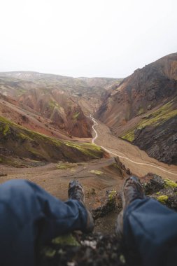 Landmannalaugar, İzlanda 'da renkli bir kanyonun kenarında oturan bir yürüyüşçü, aşağıdaki geniş ryolit manzarasını ve kıvrımlı nehri içine alır. Saf çölde ham bir an..