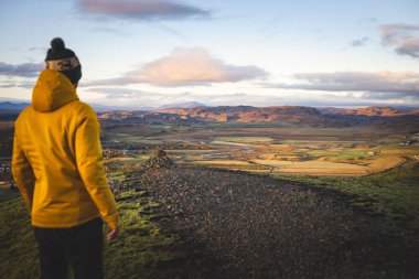 Sarı ceketli bir yürüyüşçü Hrunamannahreppur, İzlanda 'da Midfell' in tepesinde duruyor ve gün batımında altın vadi manzarasını seyrediyor. Sessiz bir düşünce ve merak anı.