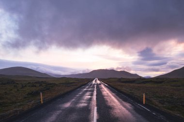 Boş yollar uzak dağlara doğru uzanır dramatik bir alacakaranlık gökyüzünün altında, mor ve pembe bulutların yansımaları ıslak asfaltta parlar..