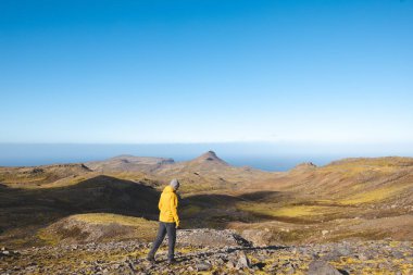 Sarı ceketli yalnız yürüyüşçü İzlanda 'daki Snaefellsnes yarımadasında, geniş volkanik dağlar ve okyanus ufkuyla çevrili parlak mavi gökyüzünün altında kayalık yolda yürüyor..