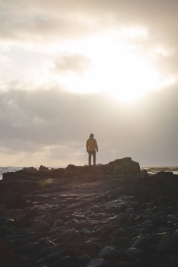 Genç gezgin, batı İzlanda 'da fırtınalı bir gün batımında kayalık bir sahil boyunca yürüyüp güneş ışığının son ışıklarının tadını çıkarıyor. Dramatik gökyüzü ve yumuşak kutup ışığı.