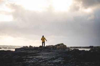 Genç gezgin, batı İzlanda 'da fırtınalı bir gün batımında kayalık bir sahil boyunca yürüyüp güneş ışığının son ışıklarının tadını çıkarıyor. Dramatik gökyüzü ve yumuşak kutup ışığı.