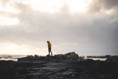 Genç gezgin, batı İzlanda 'da fırtınalı bir gün batımında kayalık bir sahil boyunca yürüyüp güneş ışığının son ışıklarının tadını çıkarıyor. Dramatik gökyüzü ve yumuşak kutup ışığı.
