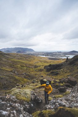 Sarı ceketli yürüyüşçü, İzlanda 'daki Snaefellsnes yarımadasındaki yosunlu kayaların ve volkanik vadinin üzerinde duruyor. Lav tarlalarının ve bulutlu gökyüzünün altındaki kıvrımlı nehrin üzerinden bakıyor..