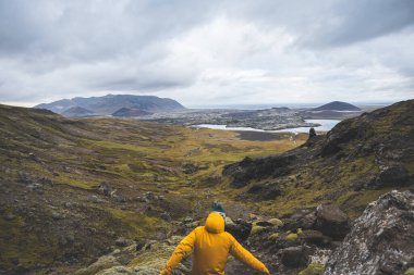 Sarı ceketli yürüyüşçü, İzlanda 'daki Snaefellsnes yarımadasındaki yosunlu kayaların ve volkanik vadinin üzerinde duruyor. Lav tarlalarının ve bulutlu gökyüzünün altındaki kıvrımlı nehrin üzerinden bakıyor..