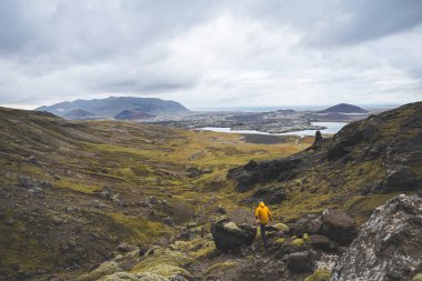 Sarı ceketli yürüyüşçü, İzlanda 'daki Snaefellsnes yarımadasındaki yosunlu kayaların ve volkanik vadinin üzerinde duruyor. Lav tarlalarının ve bulutlu gökyüzünün altındaki kıvrımlı nehrin üzerinden bakıyor..