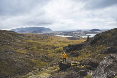 Sarı ceketli yürüyüşçü, İzlanda 'daki Snaefellsnes yarımadasındaki yosunlu kayaların ve volkanik vadinin üzerinde duruyor. Lav tarlalarının ve bulutlu gökyüzünün altındaki kıvrımlı nehrin üzerinden bakıyor..