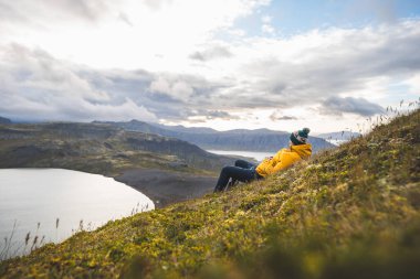 Sarı ceketli gezgin, İzlanda 'daki Snaefellsnes yarımadasındaki The Horn' un çimenli yamaçlarında dinleniyor. Huzurlu göllere, uçurumlara ve uzak dağlara bakıyor. Dağın tepesinde serinliyor.