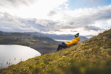 Sarı ceketli gezgin, İzlanda 'daki Snaefellsnes yarımadasındaki The Horn' un çimenli yamaçlarında dinleniyor. Huzurlu göllere, uçurumlara ve uzak dağlara bakıyor. Dağın tepesinde serinliyor.