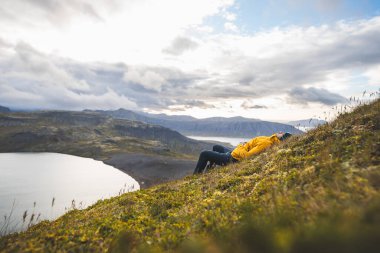 Sarı ceketli gezgin, İzlanda 'daki Snaefellsnes yarımadasındaki The Horn' un çimenli yamaçlarında dinleniyor. Huzurlu göllere, uçurumlara ve uzak dağlara bakıyor. Dağın tepesinde serinliyor.