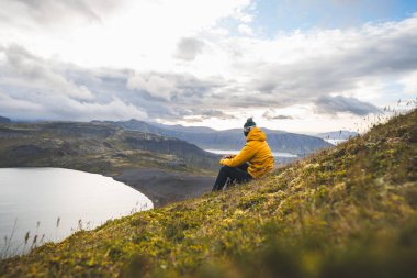 Sarı ceketli gezgin, İzlanda 'daki Snaefellsnes yarımadasındaki The Horn' un çimenli yamaçlarında dinleniyor. Huzurlu göllere, uçurumlara ve uzak dağlara bakıyor. Dağın tepesinde serinliyor.