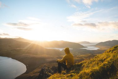 Sarı ceketli gezgin İzlanda 'daki Snaefellsnes yarımadasının tepesinde volkanik vadilerin ve aşağıdaki barışçıl göllerin üzerinden güneşin batışını izliyor..