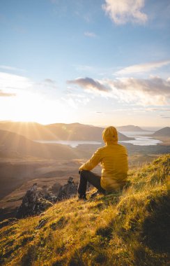 Sarı ceketli gezgin İzlanda 'daki Snaefellsnes yarımadasının tepesinde volkanik vadilerin ve aşağıdaki barışçıl göllerin üzerinden güneşin batışını izliyor..