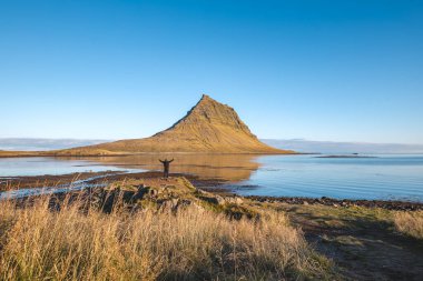 Yalnız gezgin, Snaefellsnes yarımadasındaki Kirkjufell 'e bakarak altın ışık ve yansımaların doğada huzurlu bir sabah anını şekillendirdiğini görür..