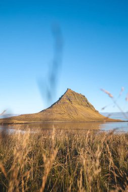 Kirkjufell 'in ikonik zirvesi Snaefellsnes yarımadasında keskin bir şekilde yükselir. Durgun bir göl ve bulutsuz gökyüzü ile altın güneş ışığıyla yıkanır ve bu mükemmel manzarayı tamamlar..