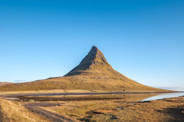 Kirkjufell 'in ikonik zirvesi Snaefellsnes yarımadasında keskin bir şekilde yükselir. Durgun bir göl ve bulutsuz gökyüzü ile altın güneş ışığıyla yıkanır ve bu mükemmel manzarayı tamamlar..