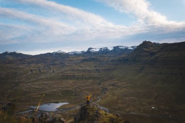 Sarı ceketli yürüyüşçü, İzlanda 'daki Kirkjufell Dağı' nın tepesinde doğan güneşi kucaklıyor. Fiyortların, vadilerin ve altın gökyüzünün üzerinde saf bir özgürlük anı yakalıyor..