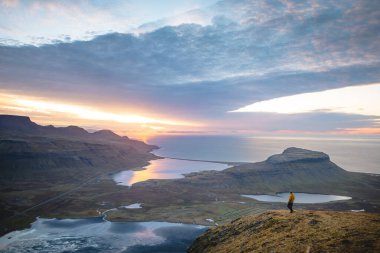 Sarı ceketli yürüyüşçü, İzlanda 'daki Kirkjufell Dağı' nın tepesinde doğan güneşi kucaklıyor. Fiyortların, vadilerin ve altın gökyüzünün üzerinde saf bir özgürlük anı yakalıyor..