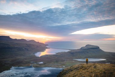 Sarı ceketli yürüyüşçü, İzlanda 'daki Kirkjufell Dağı' nın tepesinde doğan güneşi kucaklıyor. Fiyortların, vadilerin ve altın gökyüzünün üzerinde saf bir özgürlük anı yakalıyor..