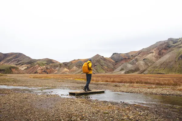 Yürüyüşçü sığ bir derenin üzerindeki ahşap bir tahtanın üzerinde duruyor İzlanda Highlands 'ın renkli rhyolite dağlarına hayran. Vahşi doğada sessiz bir an..