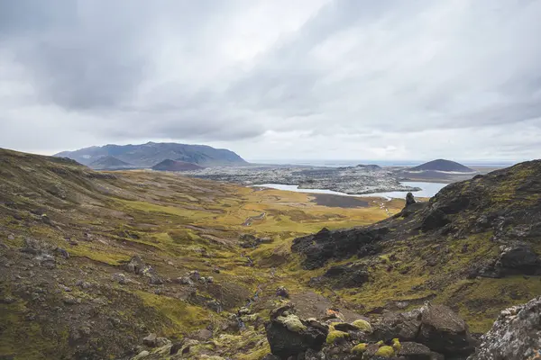 Sarı ceketli yürüyüşçü, İzlanda 'daki Snaefellsnes yarımadasındaki yosunlu kayaların ve volkanik vadinin üzerinde duruyor. Lav tarlalarının ve bulutlu gökyüzünün altındaki kıvrımlı nehrin üzerinden bakıyor..