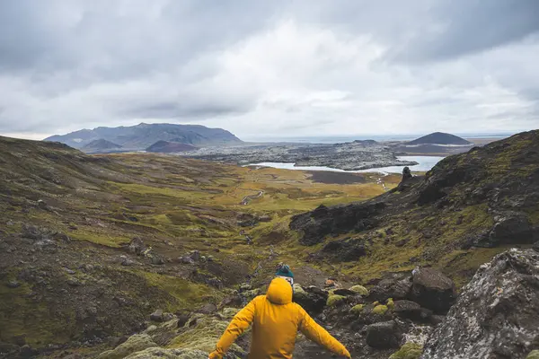 Sarı ceketli yürüyüşçü, İzlanda 'daki Snaefellsnes yarımadasındaki yosunlu kayaların ve volkanik vadinin üzerinde duruyor. Lav tarlalarının ve bulutlu gökyüzünün altındaki kıvrımlı nehrin üzerinden bakıyor..