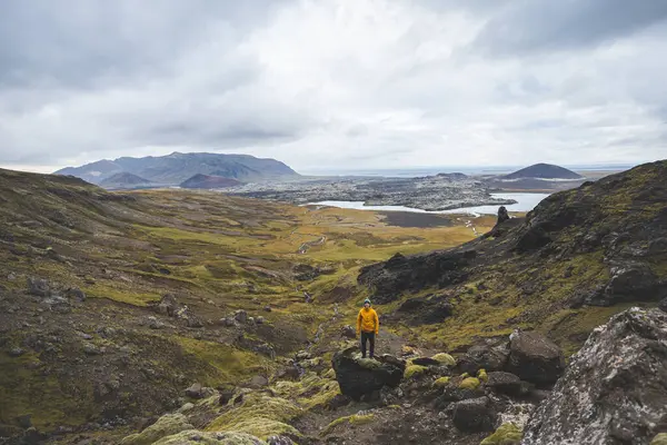 Sarı ceketli yürüyüşçü, İzlanda 'daki Snaefellsnes yarımadasındaki yosunlu kayaların ve volkanik vadinin üzerinde duruyor. Lav tarlalarının ve bulutlu gökyüzünün altındaki kıvrımlı nehrin üzerinden bakıyor..