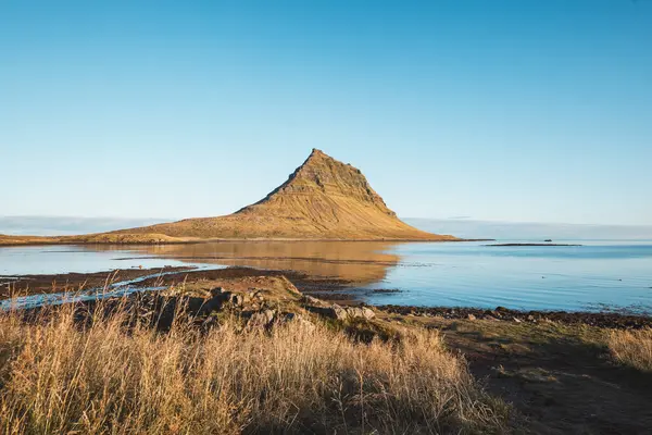 Kirkjufell 'in ikonik zirvesi Snaefellsnes yarımadasında keskin bir şekilde yükselir. Durgun bir göl ve bulutsuz gökyüzü ile altın güneş ışığıyla yıkanır ve bu mükemmel manzarayı tamamlar..