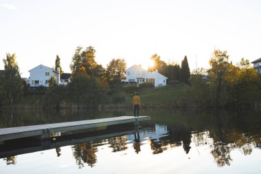 Oslo gölünde yalnız bir adam yürüyor, Holmendammen güneş büyüleyici evlerin arkasında batarken, Norveç sonbahar sihrinin huzurlu yansımasını ve dinginliğini yakalıyor..
