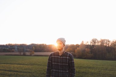 Gün batımında yeşil çayırda duran ekose gömlekli ve bereli genç adam. Altın güneş ışığı, sonbahar ağaçları ve huzurlu kırsal alan sıcak, sakin bir açık hava yaşam tarzı yaratıyor..