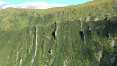 Dağların havadan görüntüsü. Dağlar, tepeler, uçurumlar, kayalar, çıkıntılar, manzara, yeşil çimenler, gökyüzü, bulutlar, doğa, işlenmemiş doğa, insansız hava aracı videosu. Dağlık bölgede hareket, 