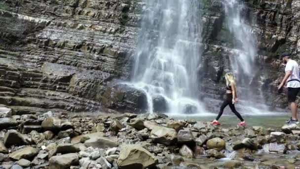 快乐的年轻夫妇仰望大瀑布 快乐的年轻夫妇仰望大瀑布 游客参观山中的瀑布 积极的生活方式人们站在美丽的瀑布上 站在瀑布边 — 图库视频影像