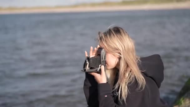 カメラ付きハッピーガール 少女は海岸の岩の上に座ってヴィンテージレトロカメラの風景に写真を撮る美しい若い旅行者です 自然へのバックパックとカメラで旅行 — ストック動画