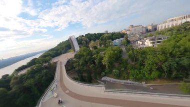 FPV drone, Reunion Arch, Kyiv: Halkın Dostluk Kemeri ve Volodymyr Hill Köprüsü. Kiev 'deki parkın ve yaya köprüsünün manzarası. Parktaki insanlar ve yeşil ağaçlar. Drone Video. Ukrayna. Avrupa