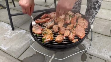 Barbeküde taze otlarla et pişiren bir kadın parti için domuz pastırmalı baharatlı sosisli piknik yapan bir kadın. Izgara domuz eti ateşte yanıyor.