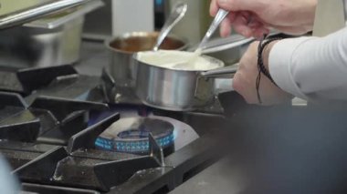 Ticari bir mutfakta çalışan profesyonel bir aşçı yemek pişirme tavaları ve diğer mutfak malzemelerinden yükselen buharlı bir yemek hazırlıyor.