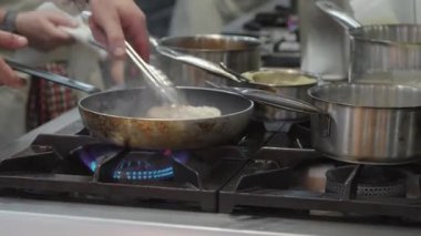 Ticari bir mutfakta çalışan profesyonel bir aşçı yemek pişirme tavaları ve diğer mutfak malzemelerinden yükselen buharlı bir yemek hazırlıyor.