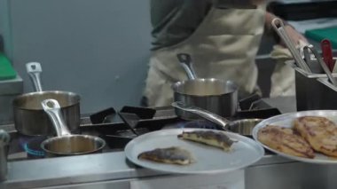 Ticari bir mutfakta çalışan profesyonel bir aşçı yemek pişirme tavaları ve diğer mutfak malzemelerinden yükselen buharlı bir yemek hazırlıyor.