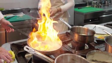 Ticari bir mutfakta çalışan profesyonel bir aşçı yemek pişirme tavaları ve diğer mutfak malzemelerinden yükselen buharlı bir yemek hazırlıyor.