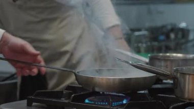 Ticari bir mutfakta çalışan profesyonel bir aşçı yemek pişirme tavaları ve diğer mutfak malzemelerinden yükselen buharlı bir yemek hazırlıyor.