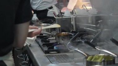 Ticari bir mutfakta çalışan profesyonel bir aşçı yemek pişirme tavaları ve diğer mutfak malzemelerinden yükselen buharlı bir yemek hazırlıyor.