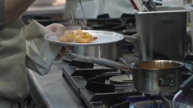 Ticari bir mutfakta çalışan profesyonel bir aşçı yemek pişirme tavaları ve diğer mutfak malzemelerinden yükselen buharlı bir yemek hazırlıyor.