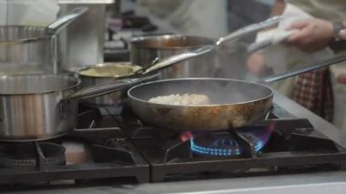 Ticari bir mutfakta çalışan profesyonel bir aşçı yemek pişirme tavaları ve diğer mutfak malzemelerinden yükselen buharlı bir yemek hazırlıyor.