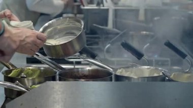 Ticari bir mutfakta çalışan profesyonel bir aşçı yemek pişirme tavaları ve diğer mutfak malzemelerinden yükselen buharlı bir yemek hazırlıyor.