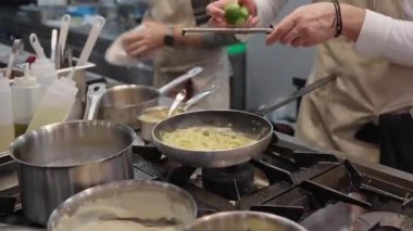 Ticari bir mutfakta çalışan profesyonel bir aşçı yemek pişirme tavaları ve diğer mutfak malzemelerinden yükselen buharlı bir yemek hazırlıyor.