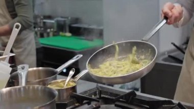 Ticari bir mutfakta çalışan profesyonel bir aşçı yemek pişirme tavaları ve diğer mutfak malzemelerinden yükselen buharlı bir yemek hazırlıyor.