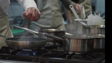 Ticari bir mutfakta çalışan profesyonel bir aşçı yemek pişirme tavaları ve diğer mutfak malzemelerinden yükselen buharlı bir yemek hazırlıyor.