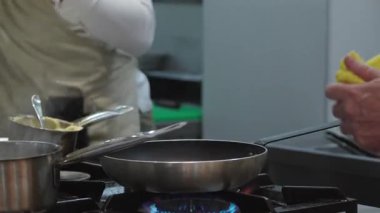 Ticari bir mutfakta çalışan profesyonel bir aşçı yemek pişirme tavaları ve diğer mutfak malzemelerinden yükselen buharlı bir yemek hazırlıyor.