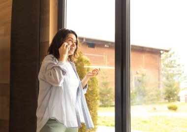 Genç bir kadın evinin panoramik penceresinin yanında telefonla konuşuyor.