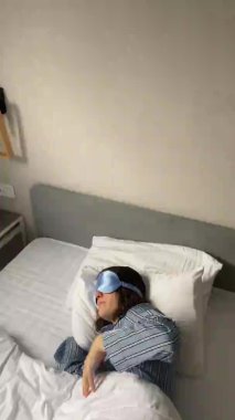 Uyku maskeli genç bir kadın yatakta uyuyor ve battaniyeyi düzeltiyor.